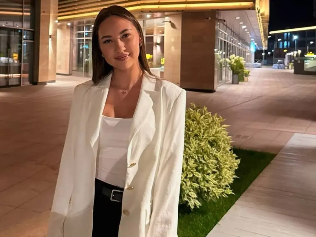 KaranAreas is wearing a white blazer and white top, posing outdoors at night near a building.