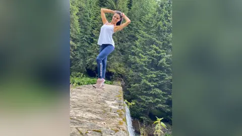 Smiling woman in white tank top and blue leggings posing on a stone ledge near a lush green forest waterfall.