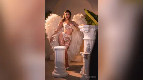 HillaryLewis poses in white lace lingerie and large white angel wings, standing next to a white classical pillar.