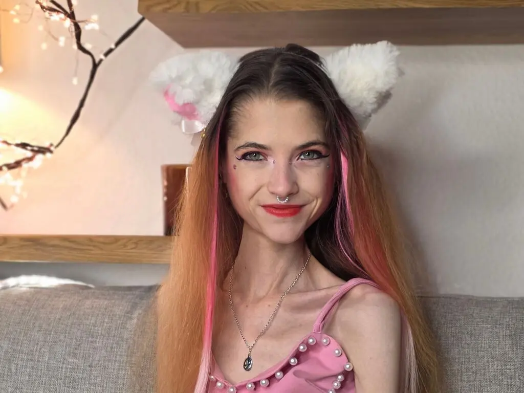 Smiling young woman with pink hair and cat ears wearing a pink strap top in a cozy indoor setting.
