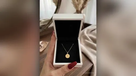 AlienaMoore — AlienaMoore holds an open white jewelry box containing a gold circular pendant necklace.
