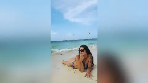 Relaxed woman in a blue bikini and sunglasses lounging on a sunny tropical beach by the turquoise ocean.