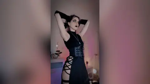 LorenLoiss poses with her arms raised, wearing a black corset and a black top with dark lipstick and heavy eye makeup.