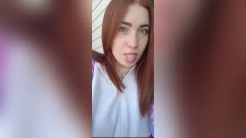 Playful redhead woman in a white hoodie sticking her tongue out against a neutral background.
