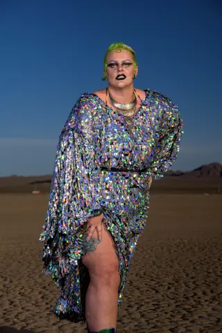 Woman in shimmering iridescent sequin dress posing confidently on a desert beach under a clear blue sky.