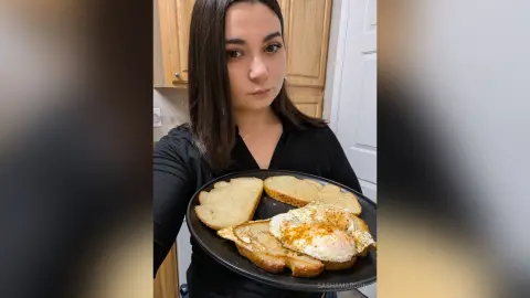 SashaMaroon holds a black plate with two pieces of toast and two fried eggs in a kitchen setting.