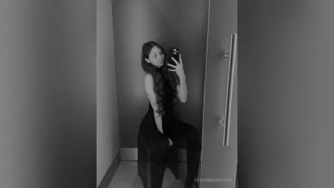 Young woman in a black bodysuit taking a moody black and white mirror selfie in a modern minimalist hallway.