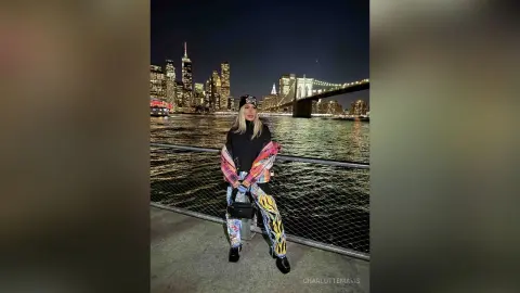 Stylish woman in patterned pants and black top posing by the river with NYC skyline at night.