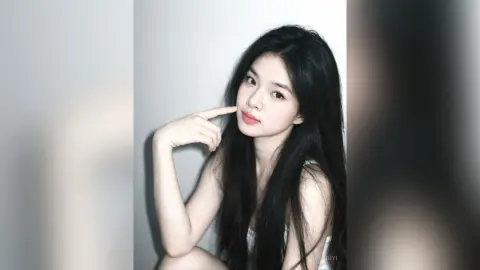 Young woman with long black hair in a white top posing with a playful expression against a plain background.