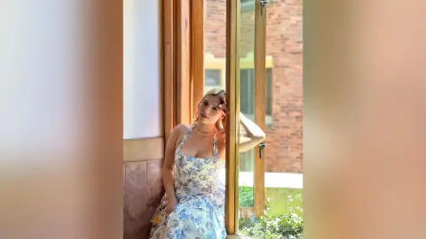 Young woman in a floral summer dress standing by a sunlit window in a bright, airy room.