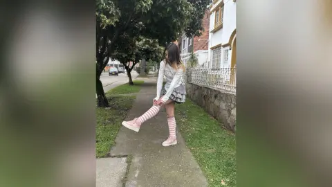 MielVerane stands on a sidewalk wearing a white long-sleeve top, a grey skirt, and pink and white striped thigh-high socks with pink boots.