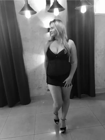 Smiling blonde woman in a black lace camisole outfit standing in a moody, dimly lit room with pendant lights.