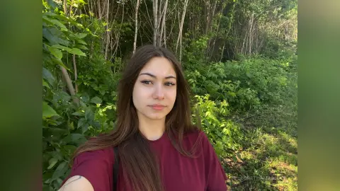 DorethaMadlock poses outdoors against a background of green leaves and trees, wearing a maroon t-shirt with her long brown hair down.