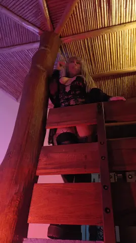 Blonde woman in black gothic outfit standing on a wooden balcony under a rustic bamboo ceiling with pink lighting.