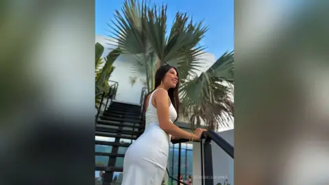 Smiling woman in a white summer dress standing on a balcony with palm trees under a clear blue sky.