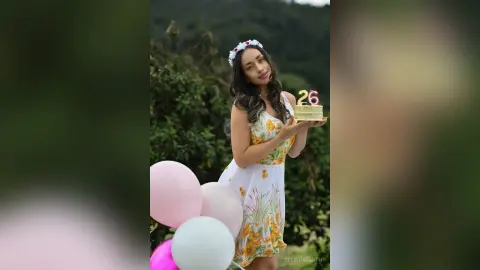 Smiling young woman in a floral sundress holding a birthday cake with a 26 candle in a lush green garden.