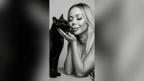 Glamorous woman in sparkling jewelry cuddling a black cat in a moody black and white portrait.