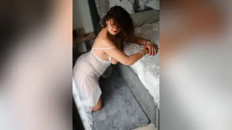 katyaros poses leaning over a bed in a sheer white chemise, showing her bare back and shoulders with her dark curly hair falling forward.