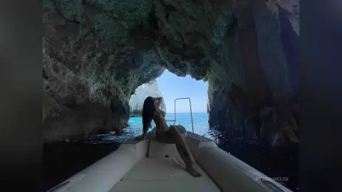 Woman in a bikini relaxing on a white boat inside a scenic sea cave with turquoise water.
