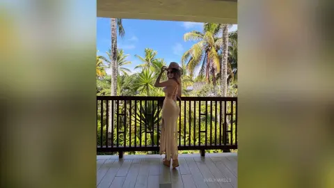 Stylish woman in a beige maxi dress and sun hat posing on a tropical balcony overlooking palm trees.