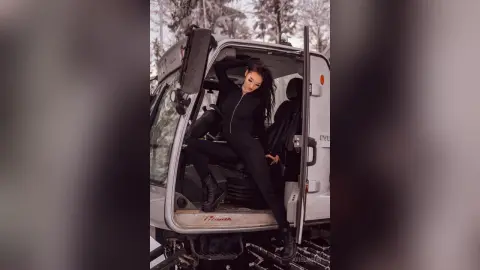 Stylish woman in a black jumpsuit leaning against a silver truck in a snowy winter landscape.