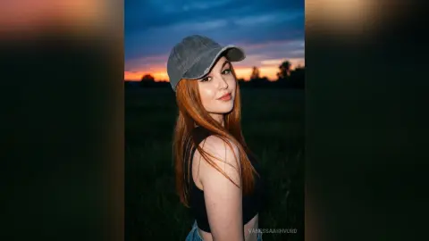 Redheaded woman in a black tank top and grey baseball cap smiling during a golden hour sunset in a grassy field.