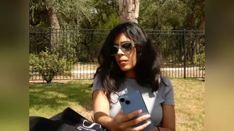 Young woman in a grey t-shirt and sunglasses relaxing on a sunny lawn near a black metal fence.