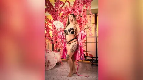 Blonde woman in black lace lingerie and pink floral kimono holding a fan in a cherry blossom setting.