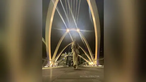 Stylish man in a beige coat walking across a modern illuminated bridge at night under dramatic architectural lights.
