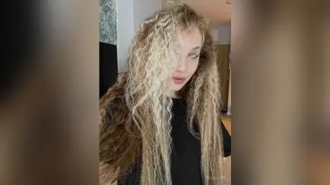 Young woman with long curly blonde hair wearing a black top in a bright indoor setting.