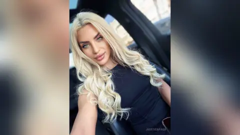 JessycaPage is a blonde woman in a black top posing for a selfie inside a car.