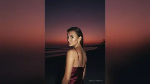 Elegant woman in a burgundy satin slip dress posing against a moody sunset beach backdrop.