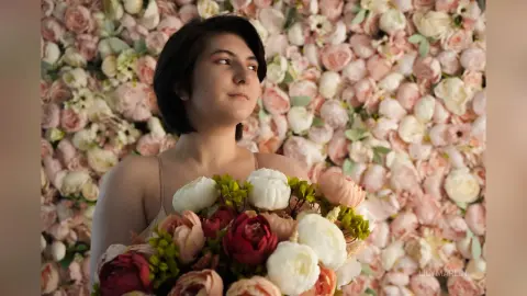 Young woman in a neutral dress holding a large bouquet of pink and white roses against a floral wall background.