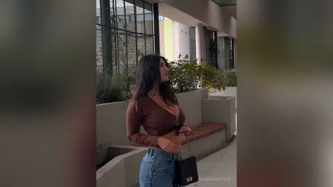 Young woman in a brown long-sleeve top and denim jeans standing outdoors in a modern urban setting.