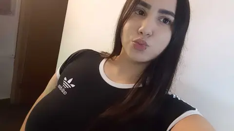 Young woman with long dark hair wearing a black Adidas t-shirt in a bright indoor setting.