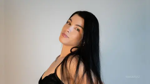 Portrait of a woman with long black hair wearing a black lace top against a minimalist white background.
