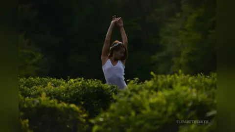 Serene woman in a white tank top stretching her arms in a lush green garden at twilight.