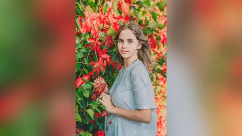 Young woman in a grey dress smiling amidst vibrant red and green autumn foliage in a peaceful garden setting.