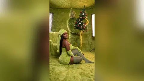 Woman in green fuzzy outfit sitting in a lime green room next to a decorated Christmas tree with a Grinch figure.
