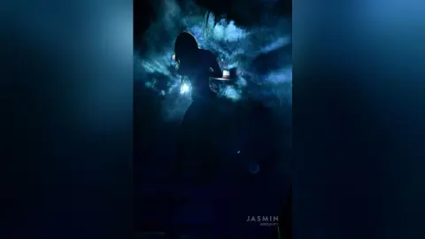 AndreaKalo stands in a dark, smoky setting, silhouetted against a blue light that highlights her body and long hair.
