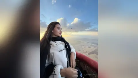 AlanaPearce poses outdoors against a blue sky with clouds, wearing a white top and a black scarf or wrap over her shoulders.