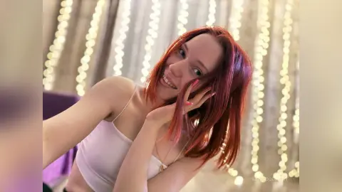 Smiling redhead woman in a white tank top posing against a dreamy background of glowing fairy lights.