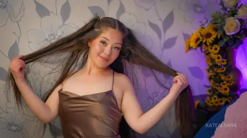KateAldana holds her long brown hair out to the sides, wearing a brown satin camisole against a floral wallpaper background.