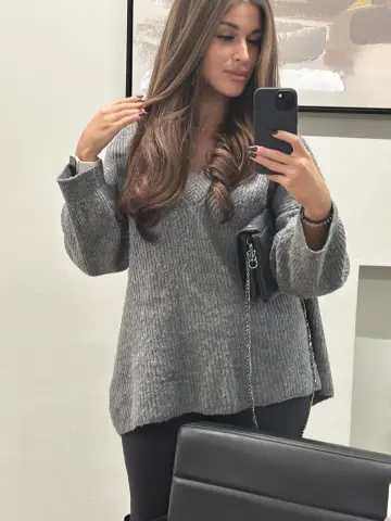 Young woman in a cozy grey oversized knit sweater posing for a mirror selfie in a bright room.