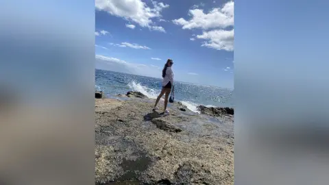 Young woman in white shirt and black shorts standing on rocky coast under a bright blue sky.