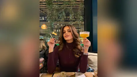 AnaMathers holds a cocktail glass in one hand and a lit cigarette in the other while wearing a dark long-sleeved top.