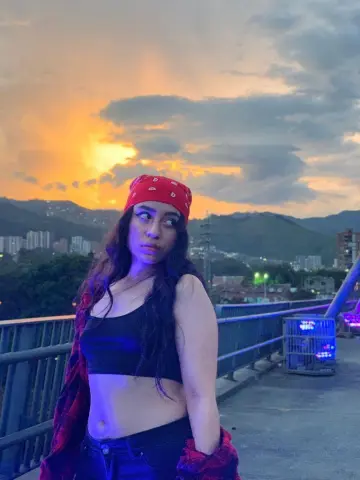 Young woman in a red bandana and black crop top posing against a dramatic sunset sky over mountain cityscapes.