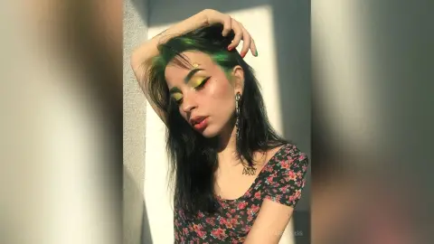 Young woman with green hair and floral top posing in sunlight against a white wall.