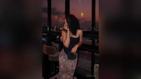 Curly-haired woman in a black tank top and patterned skirt posing by a window during a moody sunset.