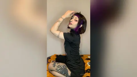 Woman with dark hair and tattoos wearing a black dress and fishnet tights in a minimalist indoor setting.